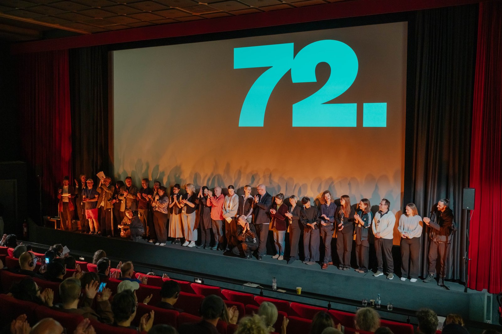 Rund 30 Leute des Kurzfilmtage-Teams stehen auf der Bühne der Lichtburg, bekommen Applaus vom Publikum und applaudieren für's Publikum.